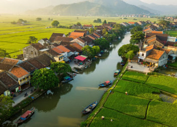 An aerial view of a traditional Vietnamese riverside village with terracotta-roofed houses, lush green rice paddies, narrow waterways with boats, and mountainous backdrop in the distance.