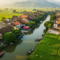An aerial view of a traditional Vietnamese riverside village with terracotta-roofed houses, lush green rice paddies, narrow waterways with boats, and mountainous backdrop in the distance.