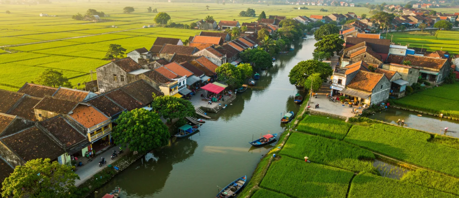 An aerial view of a traditional Vietnamese riverside village with terracotta-roofed houses, lush green rice paddies, narrow waterways with boats, and mountainous backdrop in the distance.