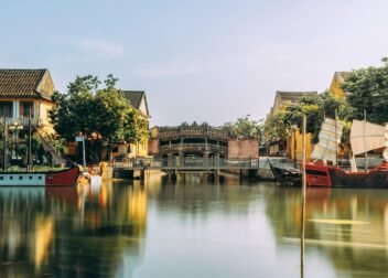 A serene waterway in Hoi An, Vietnam features traditional yellow-painted buildings, a curved wooden bridge, and colorful boats with red hulls and white sails reflected in the calm water.