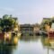 A serene waterway in Hoi An, Vietnam features traditional yellow-painted buildings, a curved wooden bridge, and colorful boats with red hulls and white sails reflected in the calm water.