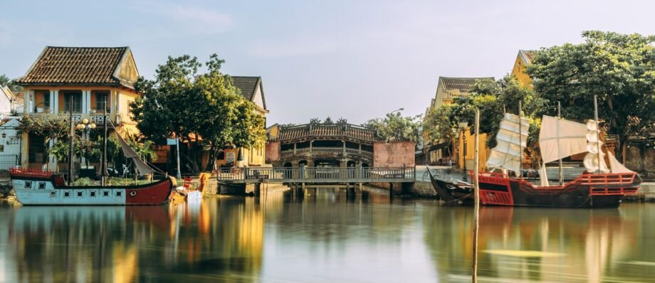 A serene waterway in Hoi An, Vietnam features traditional yellow-painted buildings, a curved wooden bridge, and colorful boats with red hulls and white sails reflected in the calm water.